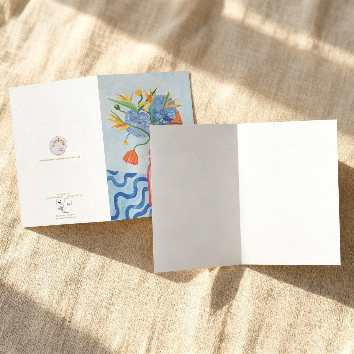 A folded white greeting card or booklet lies open on a beige linen surface with soft sunlight and shadows. The left panel shows a colorful floral illustration with blue, yellow, orange, and green tones, and the right panel is blank white.