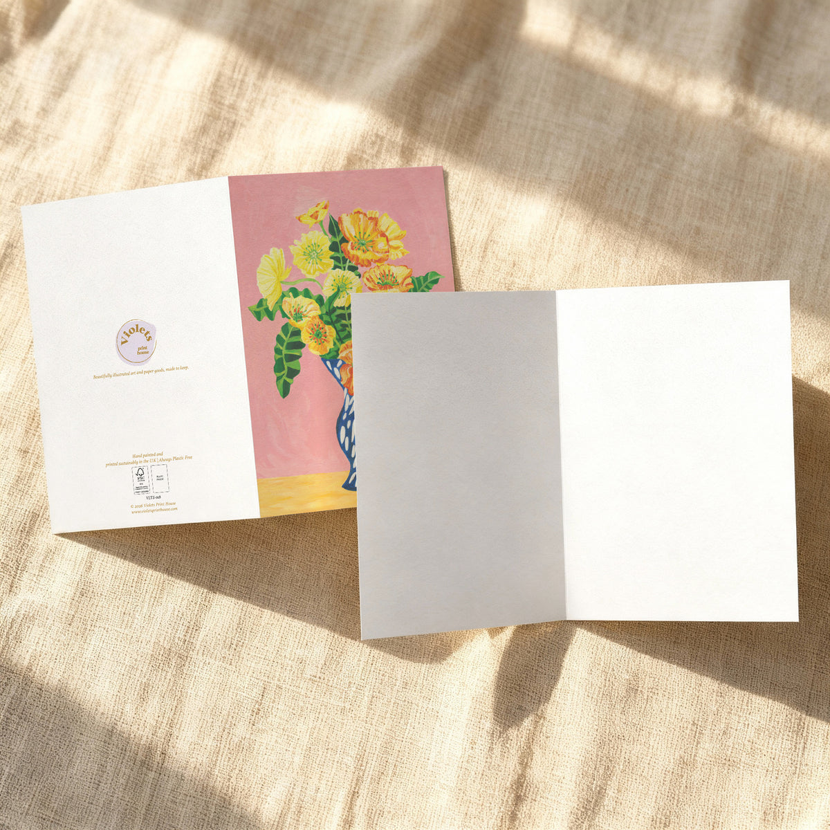 A folded greeting card or art print shown open on a textured beige fabric background with dappled sunlight. The left panel is white with a small circular logo and text, and the center panel features a colorful illustration of yellow flowers in a blue vase against a pink background.