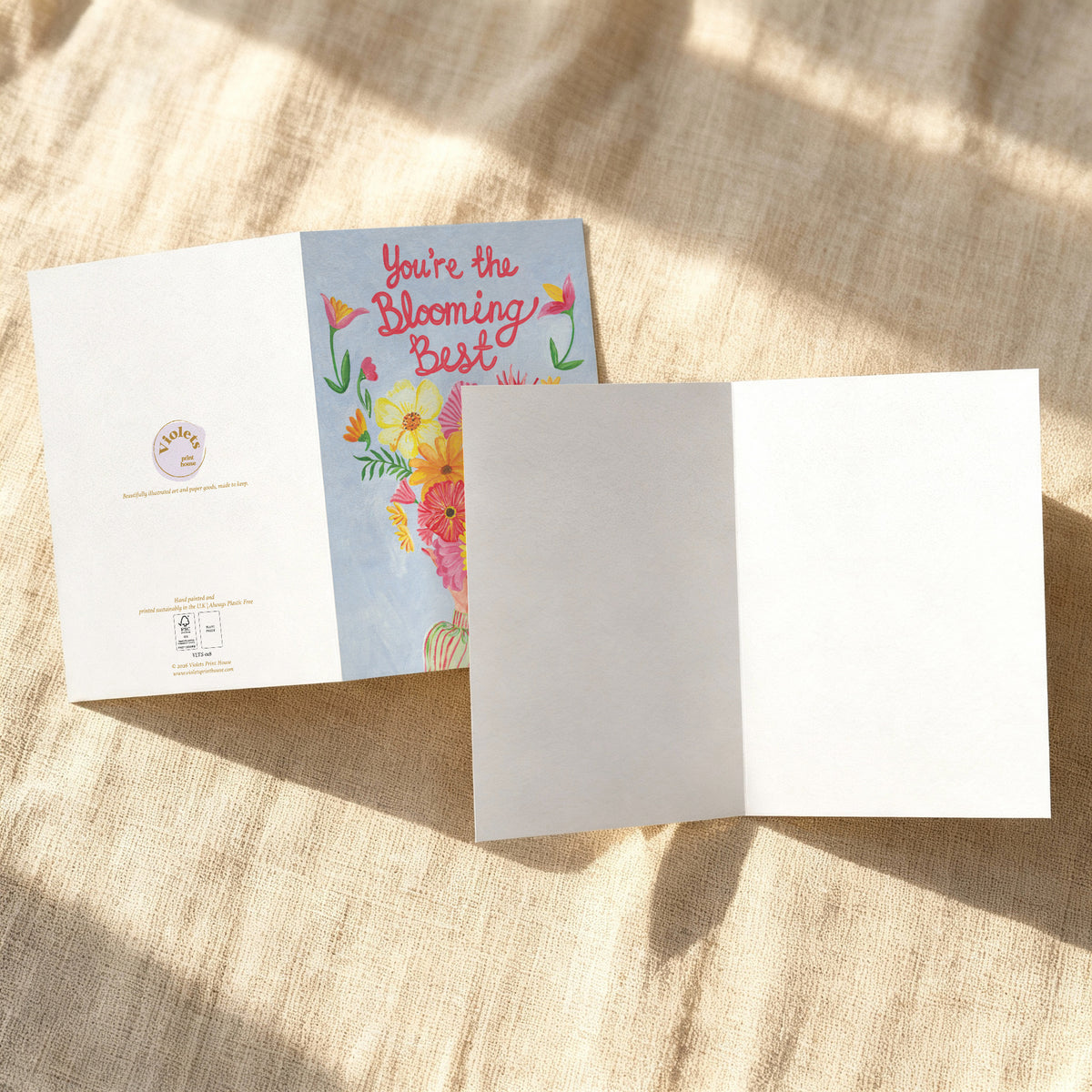An open greeting card laid on a beige fabric surface with dappled sunlight. The left panel shows a floral illustration and the text 'You're the blooming best,' while the right panel is blank white.